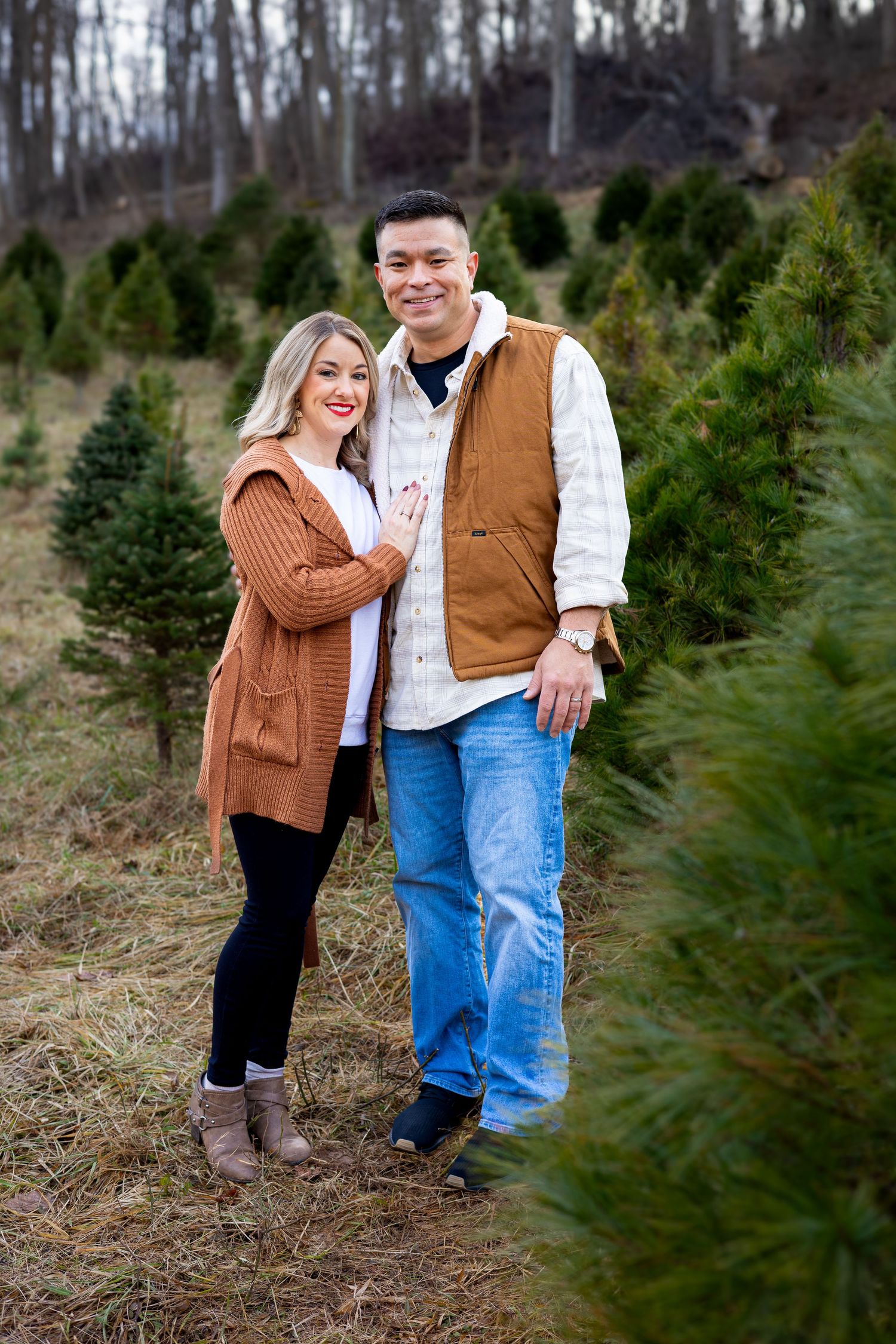 Couple in casual winter wear poses among evergreen trees at tree farm.
