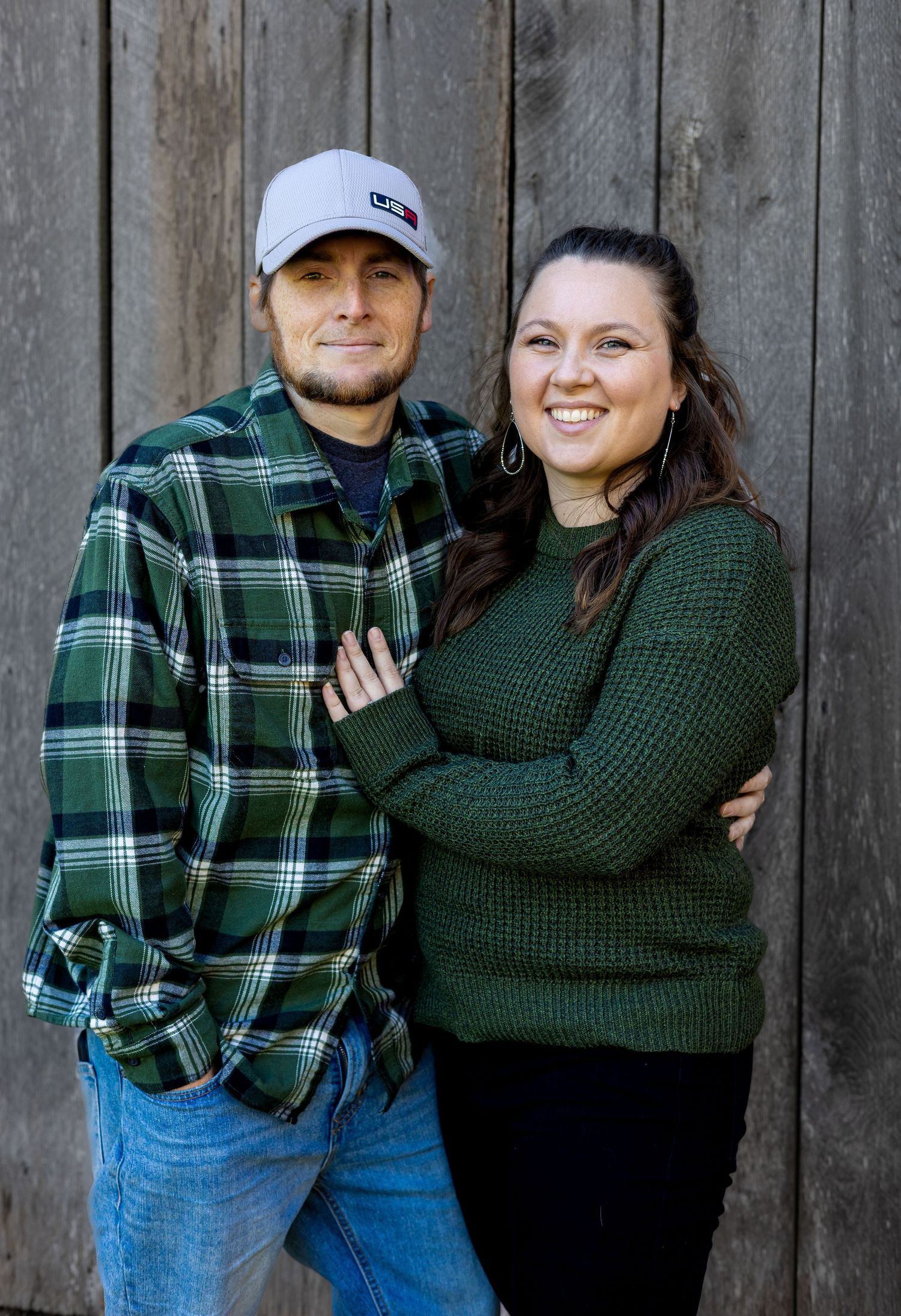 Couple in winter attire poses together against rustic wooden background.