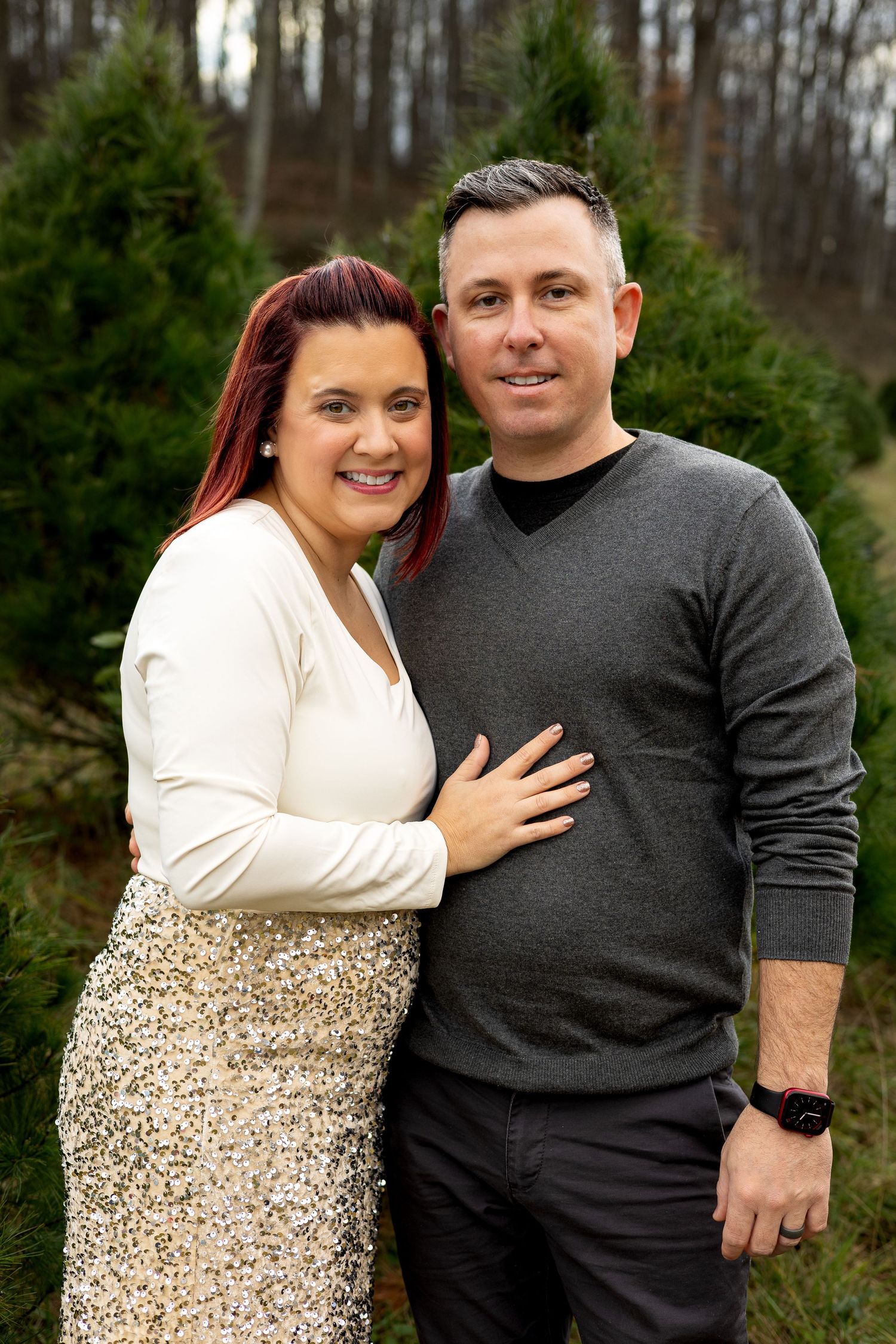 Couple poses together against evergreen trees in winter setting.