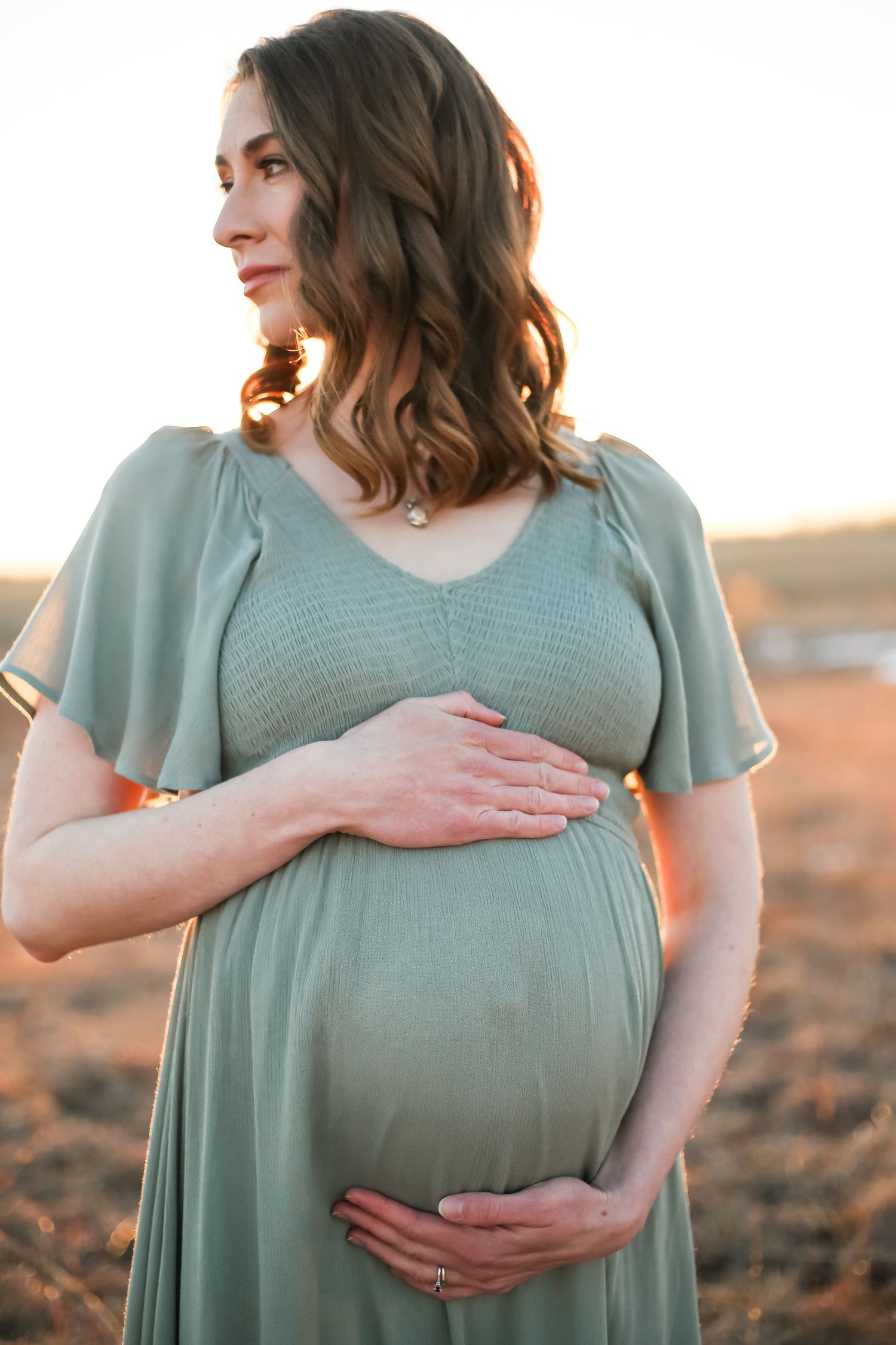 Expectant mother in sage green dress tenderly holding her baby bump during golden hour sunset outdoor photo session.