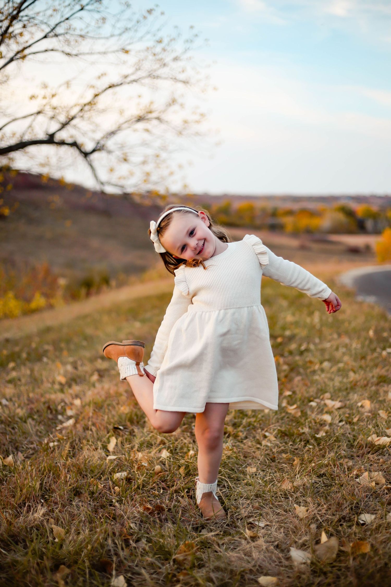A child in a white dress dances playfully on a hill during autumn with golden light and fall colors in Manhattan KS