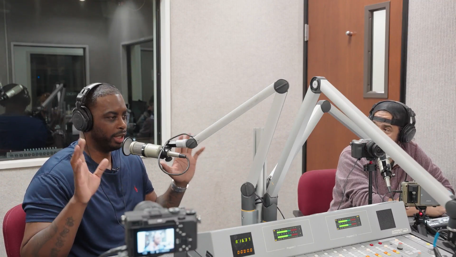Two men wearing headphones speak into microphones at a radio station broadcasting desk.