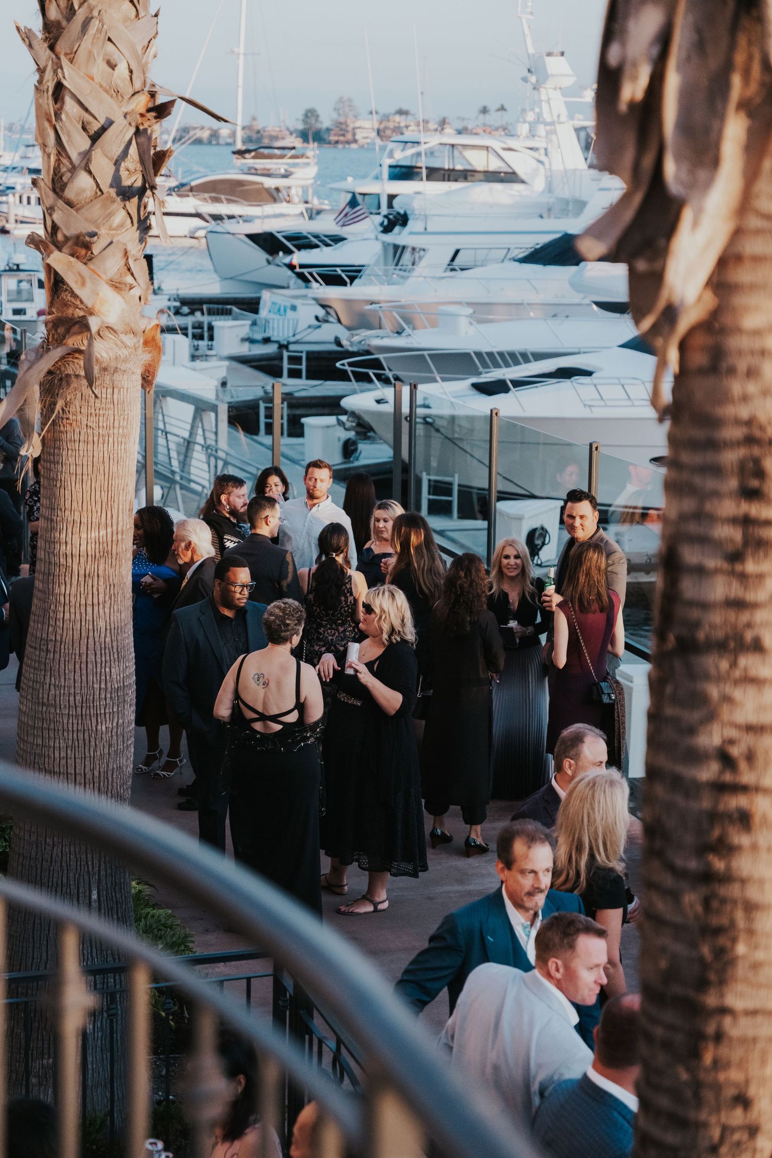 People gather for an evening social event at a marina with luxury yachts and palm trees in the background.