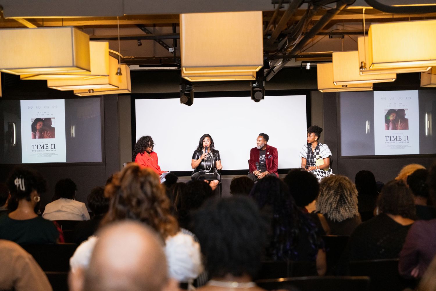 Panel discussion on stage with speakers and audience members in a dimly lit venue with modern lighting fixtures.