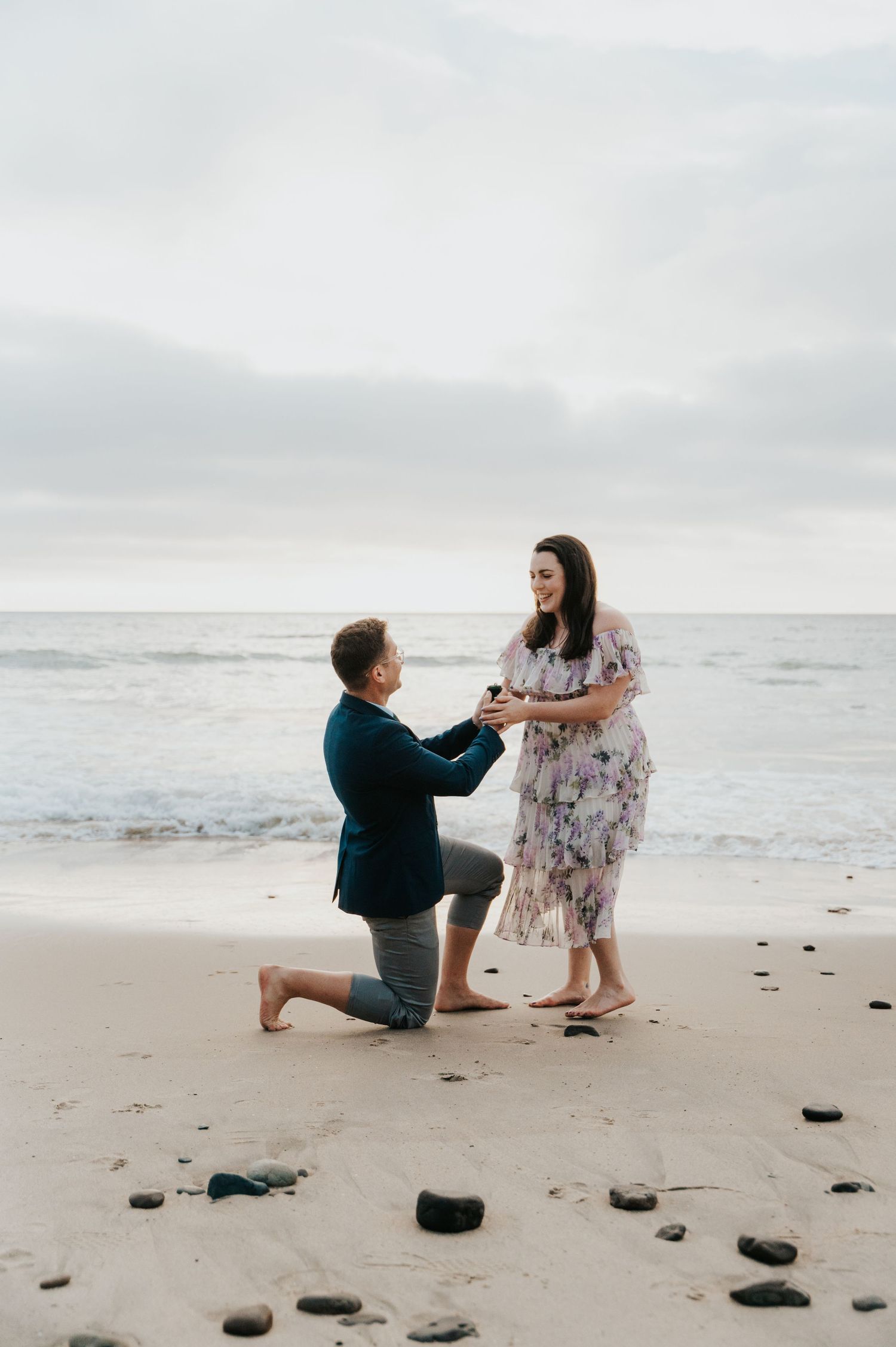 A couple shares a romantic moment during a proposal on a misty beach with scattered rocks on the sand.