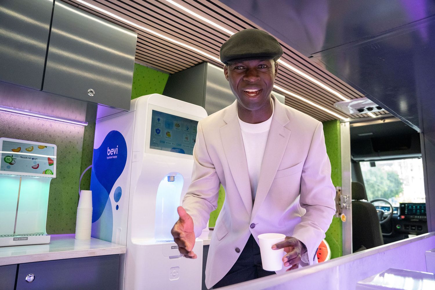Man in white blazer and cap smiling while holding a cup at a modern green and white food kiosk.