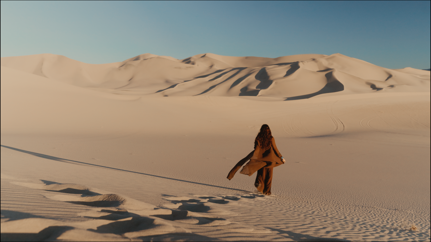A lone figure in flowing robes walks across vast golden sand dunes under a clear blue sky in a desert landscape.