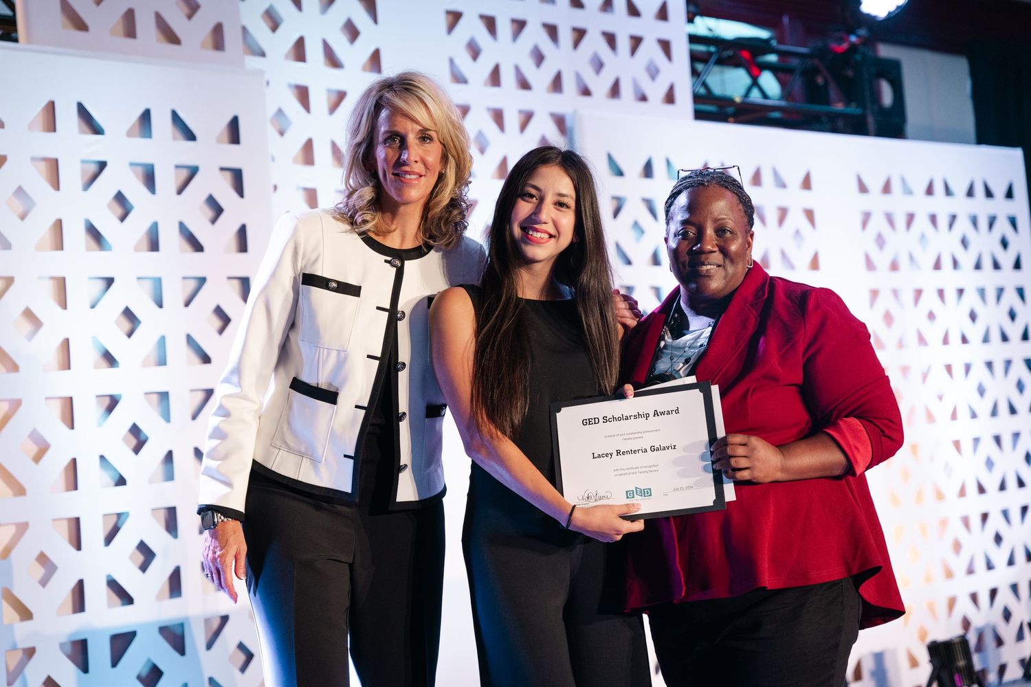 Three women smile together at an awards ceremony, with two presenting a scholarship certificate to the recipient.