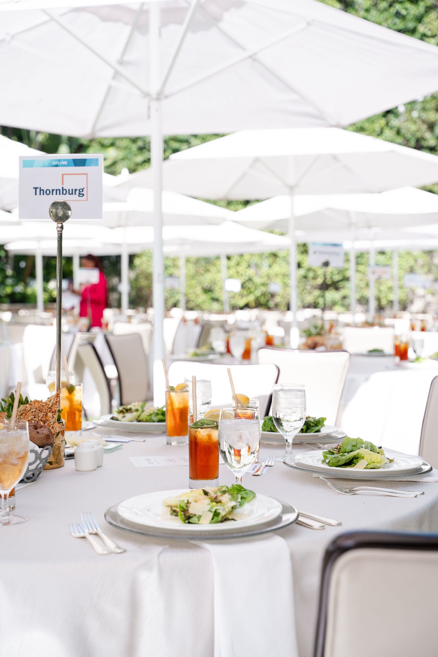 Elegant wedding reception tables set with white linens, salad plates, and orange cocktails under outdoor white umbrellas.