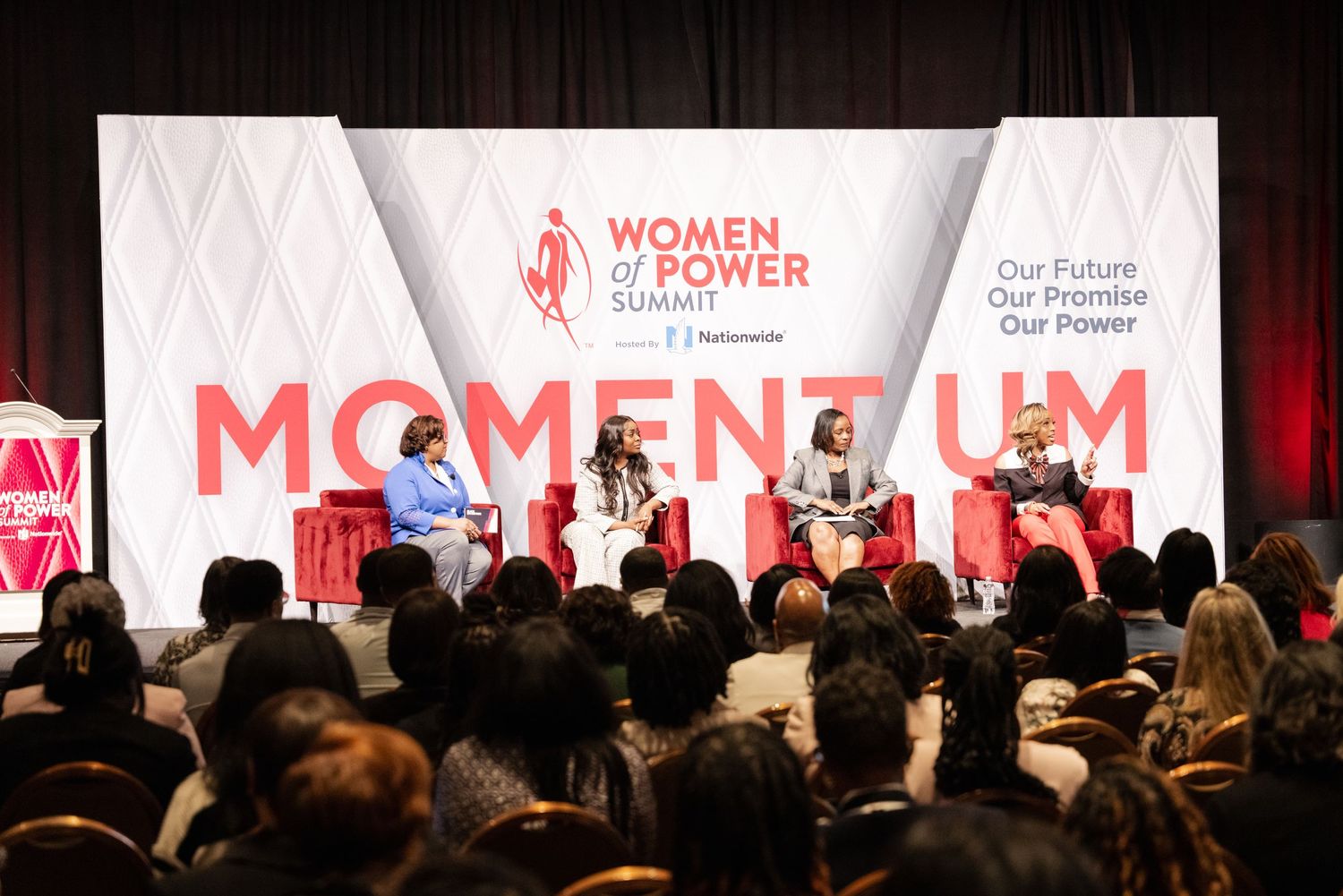 A large conference room filled with attendees watching a presentation with a red and white Momentum banner on stage.