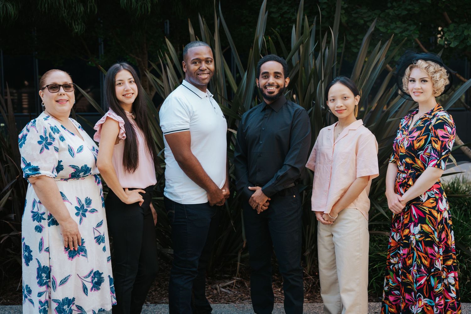 Six diverse young professionals posing together outdoors in front of tropical plants for a group photo.