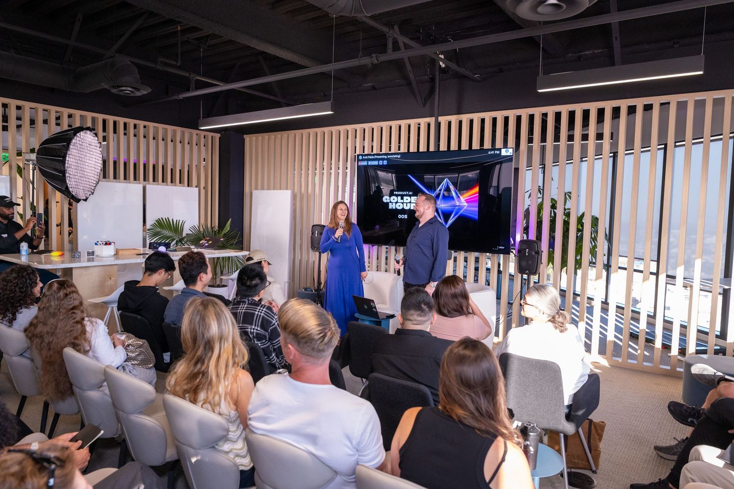 Two presenters speak on stage before an audience at a modern corporate event with a large display screen.