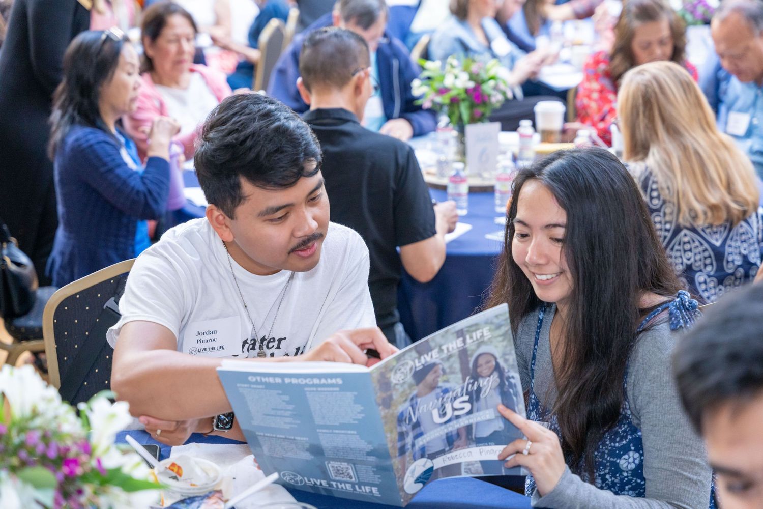 Two people sit together at an outdoor dining event looking at a printed publication while others gather in background.