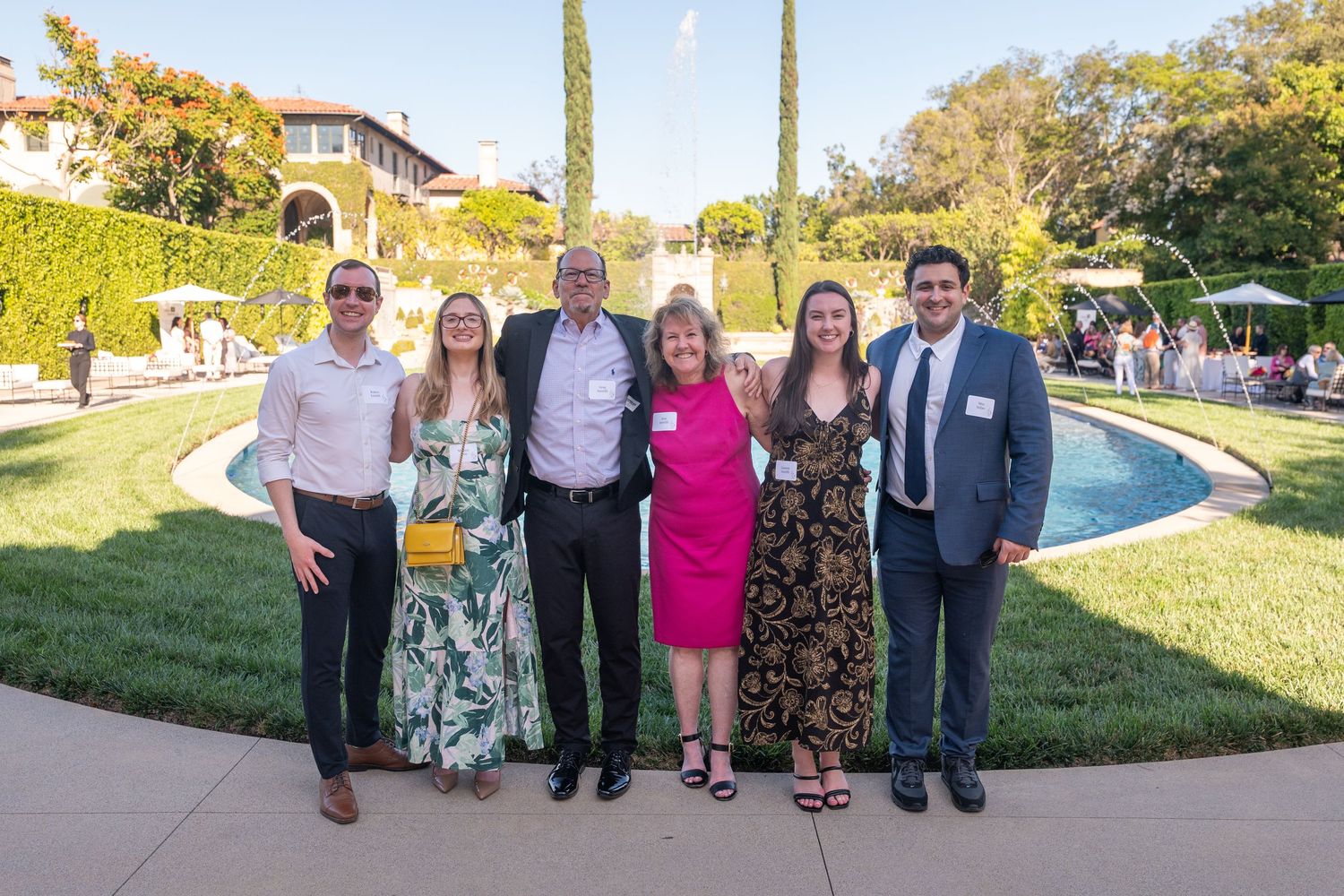 Group of six people posing outdoors at an elegant garden event with a fountain and manicured lawn.