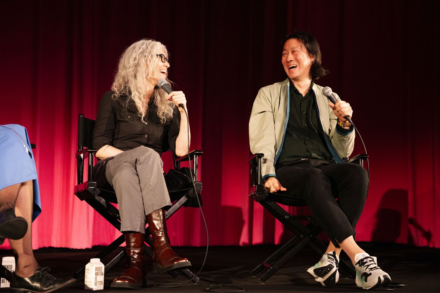 Two people sit in chairs on stage having an animated discussion in front of a red curtain backdrop.