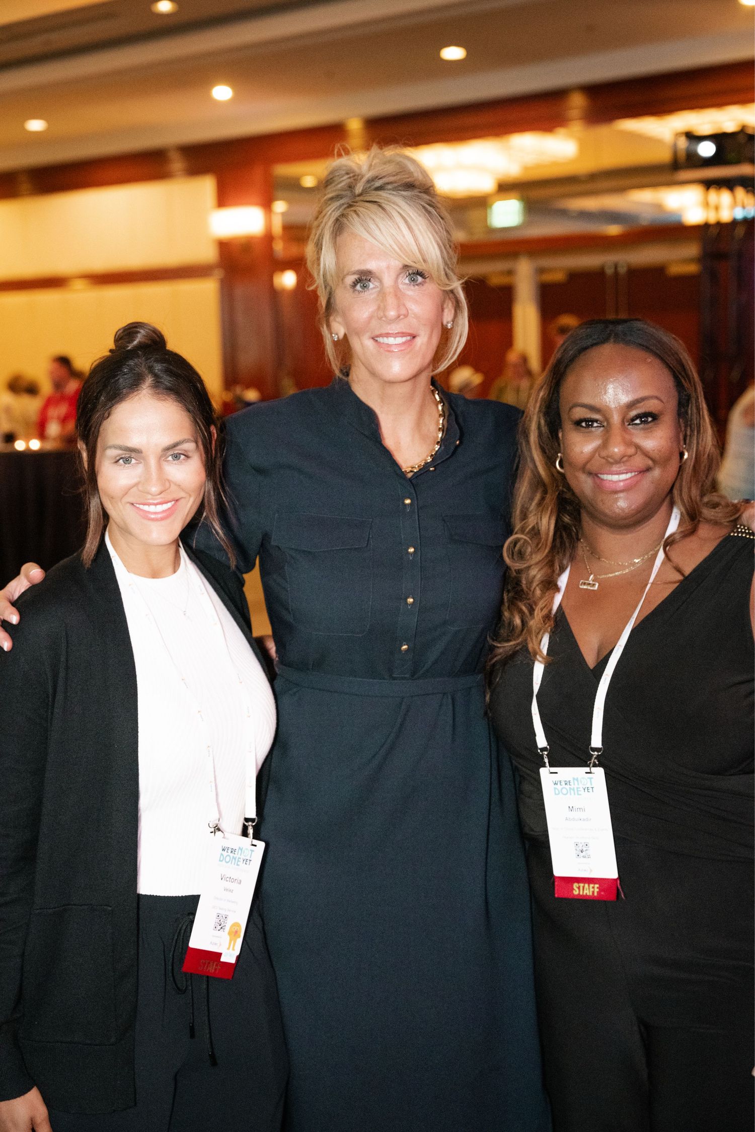 Three women wearing business attire pose together at a professional event, wearing name badges and smiling warmly.