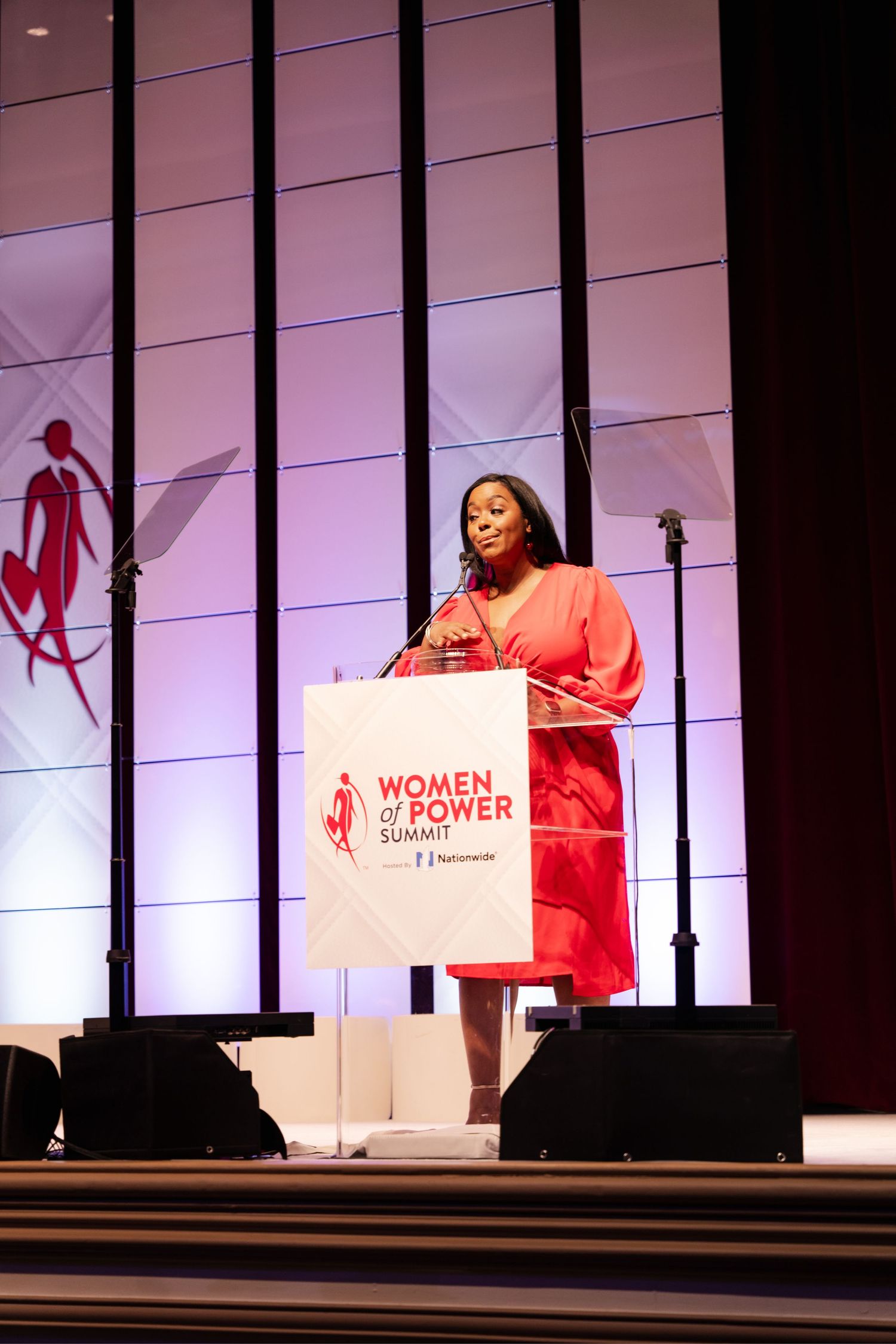 A speaker stands at a podium during a Women in Power event with a purple-lit stage backdrop.