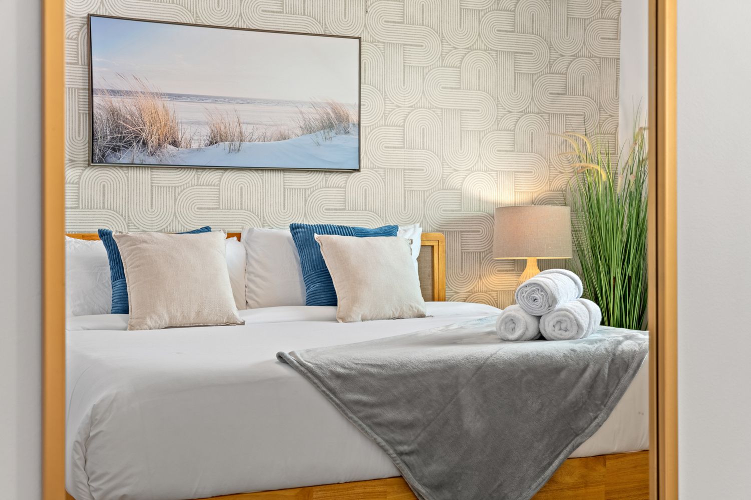 Modern bedroom interior with white bedding, blue accent pillows, wooden bed frame and coastal artwork above the headboard.
