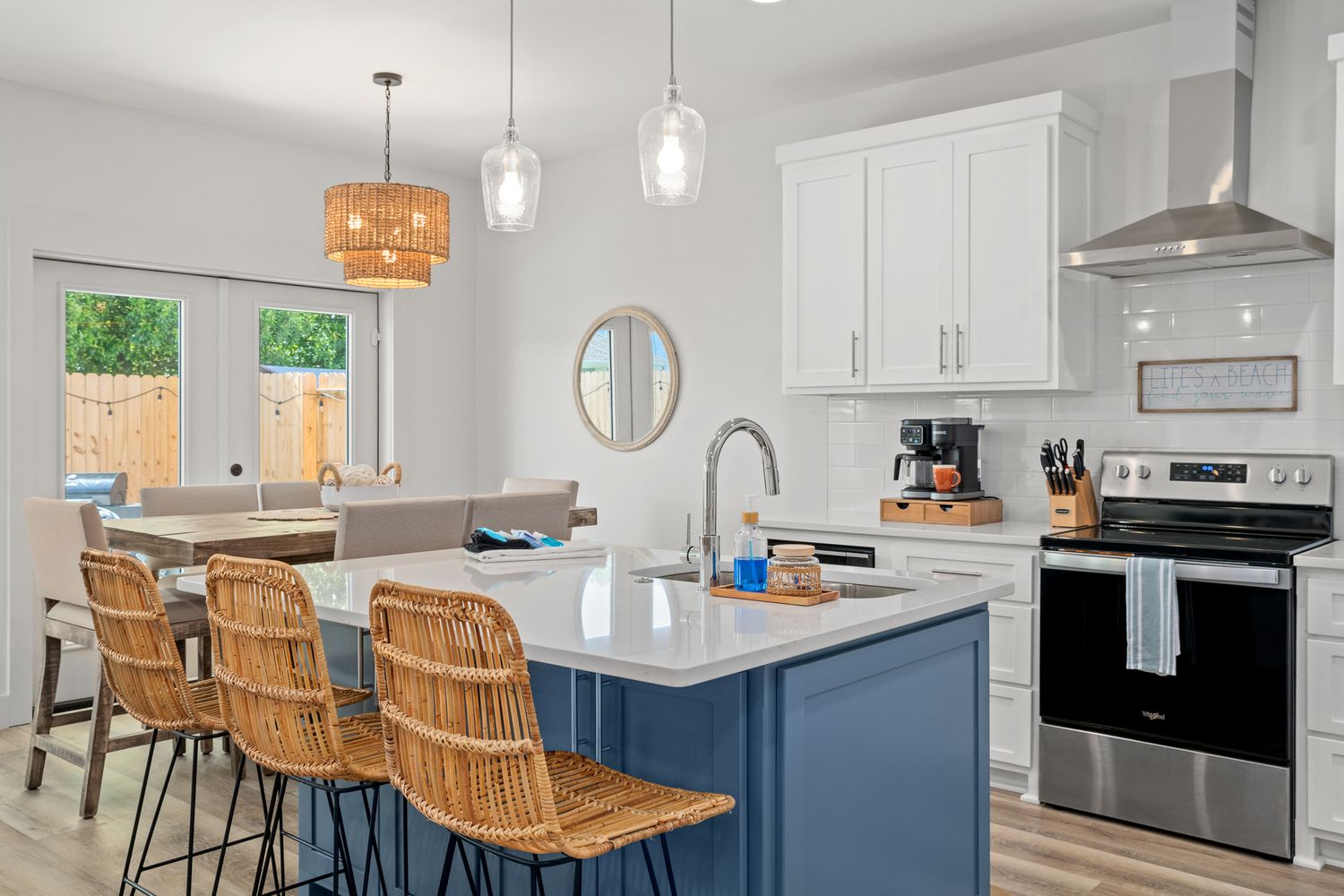 Modern kitchen with navy blue island, wicker barstools, white cabinets and pendant lighting over quartz countertops.