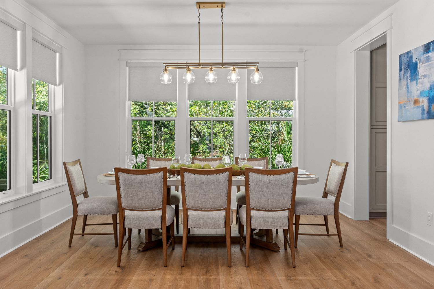 Modern dining room with hardwood floors, white walls, large windows, and a contemporary gold light fixture above wood table.