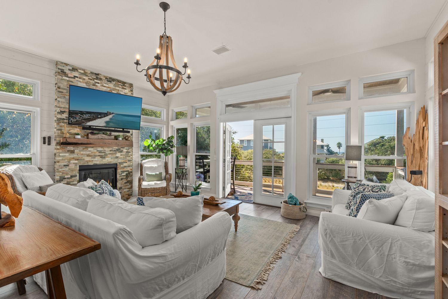 Bright coastal living room with stone fireplace, chandelier, white slip-covered furniture and large windows overlooking water views.