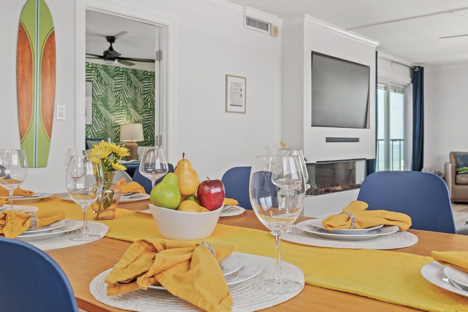 Modern dining room with yellow placemats, surfboard decor, and blue chairs around a white table set for a meal.