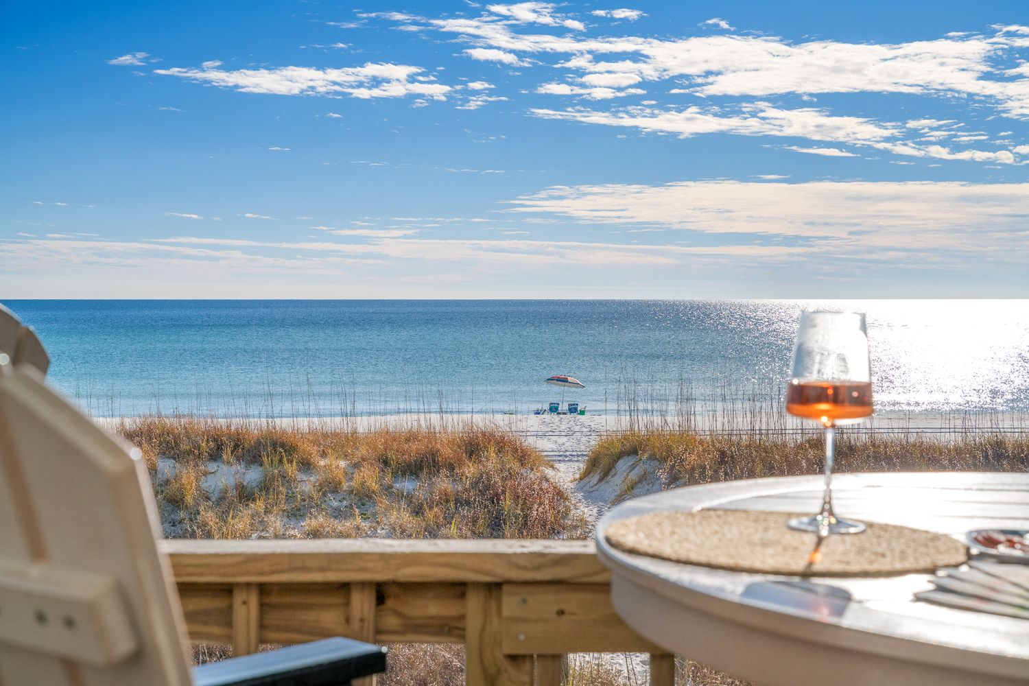 Glass of beer on wooden deck railing overlooking sunny beach and ocean with bright blue sky and white clouds.