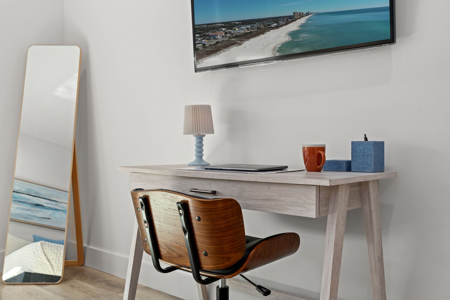 Modern home office setup with wooden desk, vintage chair, tall mirror, and coastal artwork on white walls.