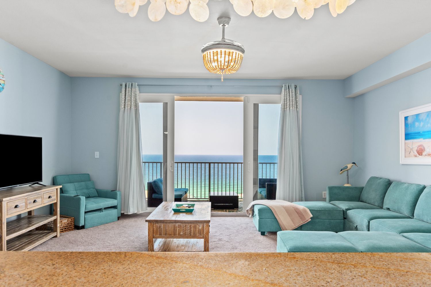 Bright coastal living room with turquoise furniture and large oceanfront balcony window offering stunning beach views.
