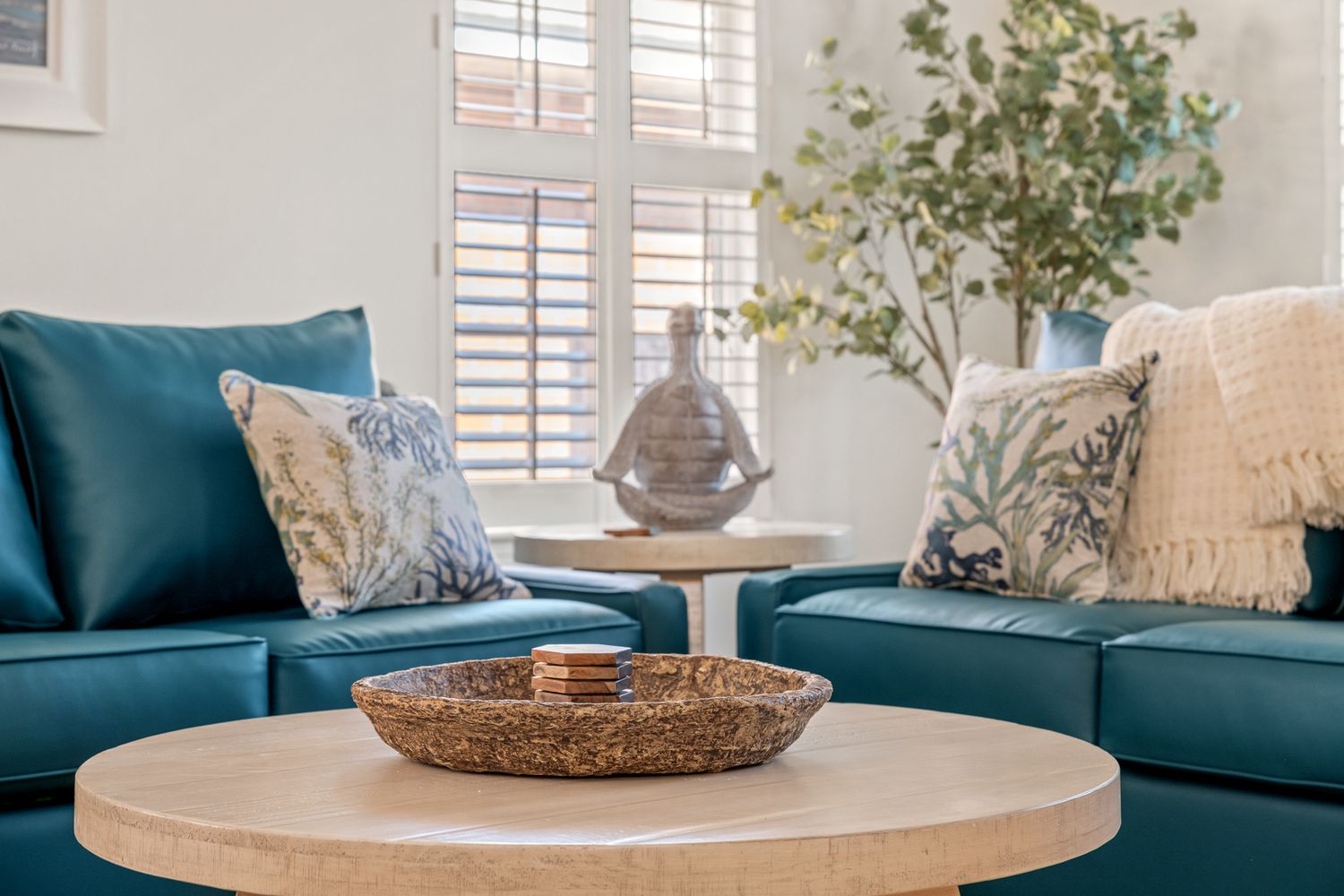 Contemporary living room features teal blue sofas with decorative pillows and a round wooden coffee table with woven bowl decor.