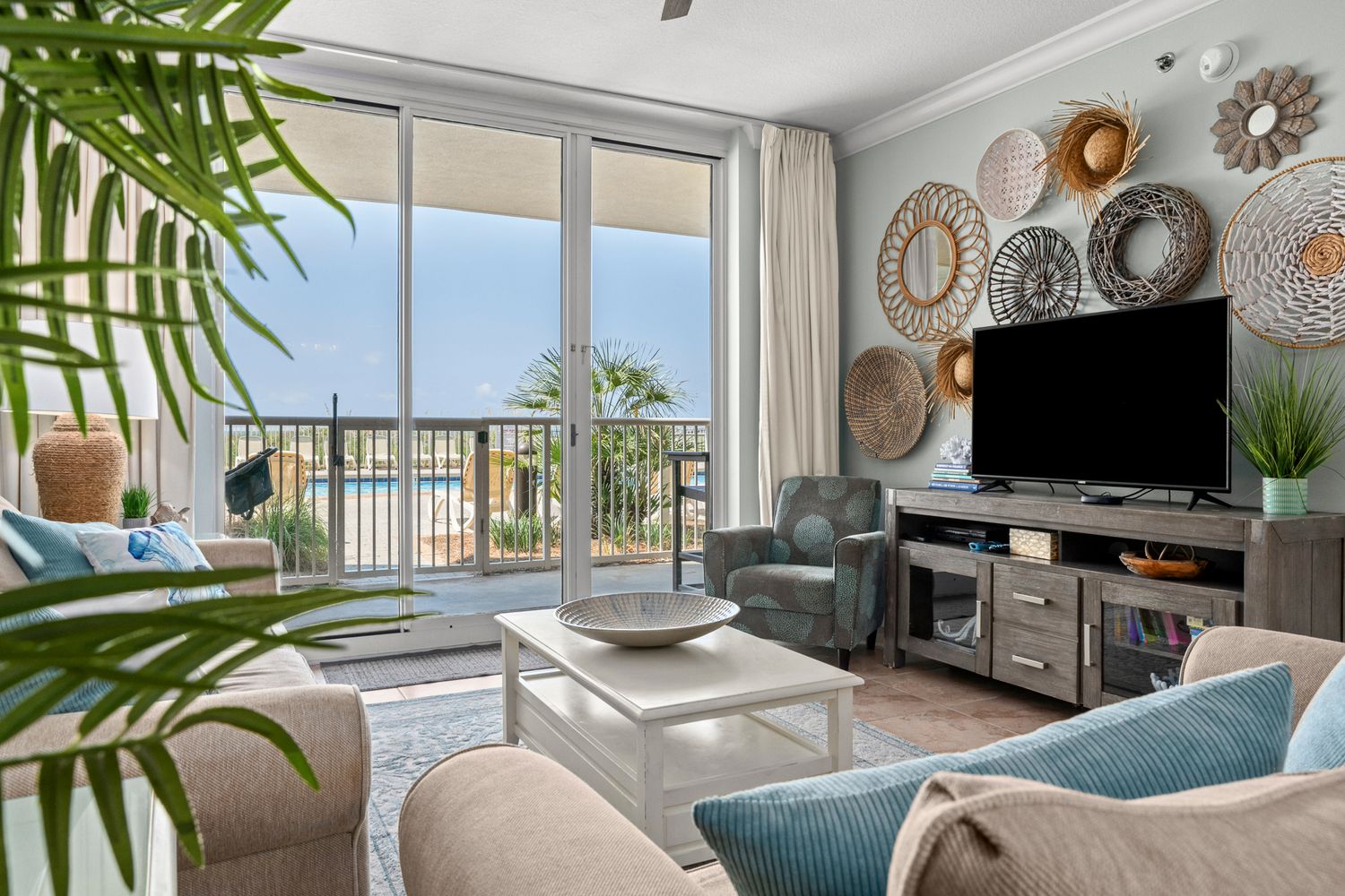 Coastal living room with ocean view balcony features beige and blue decor, wall-mounted TV, and decorative plate wall art.