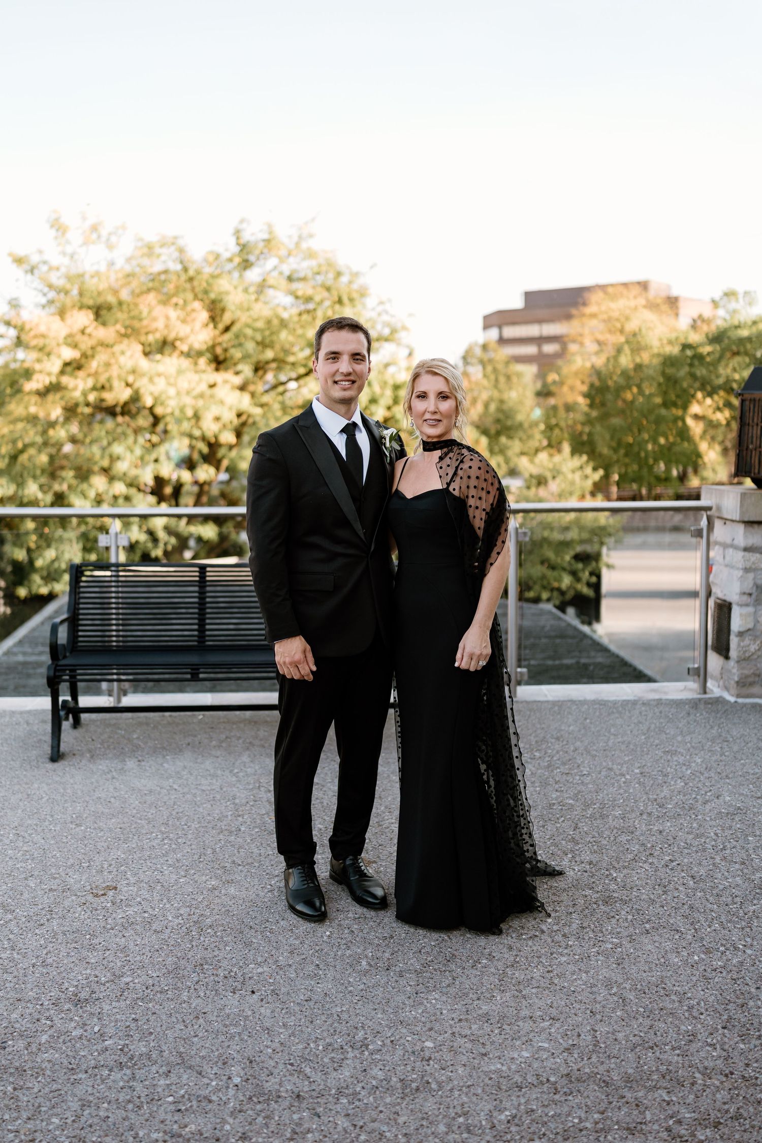 Couple in formal attire poses together on gravel path with trees and buildings in background at sunset.