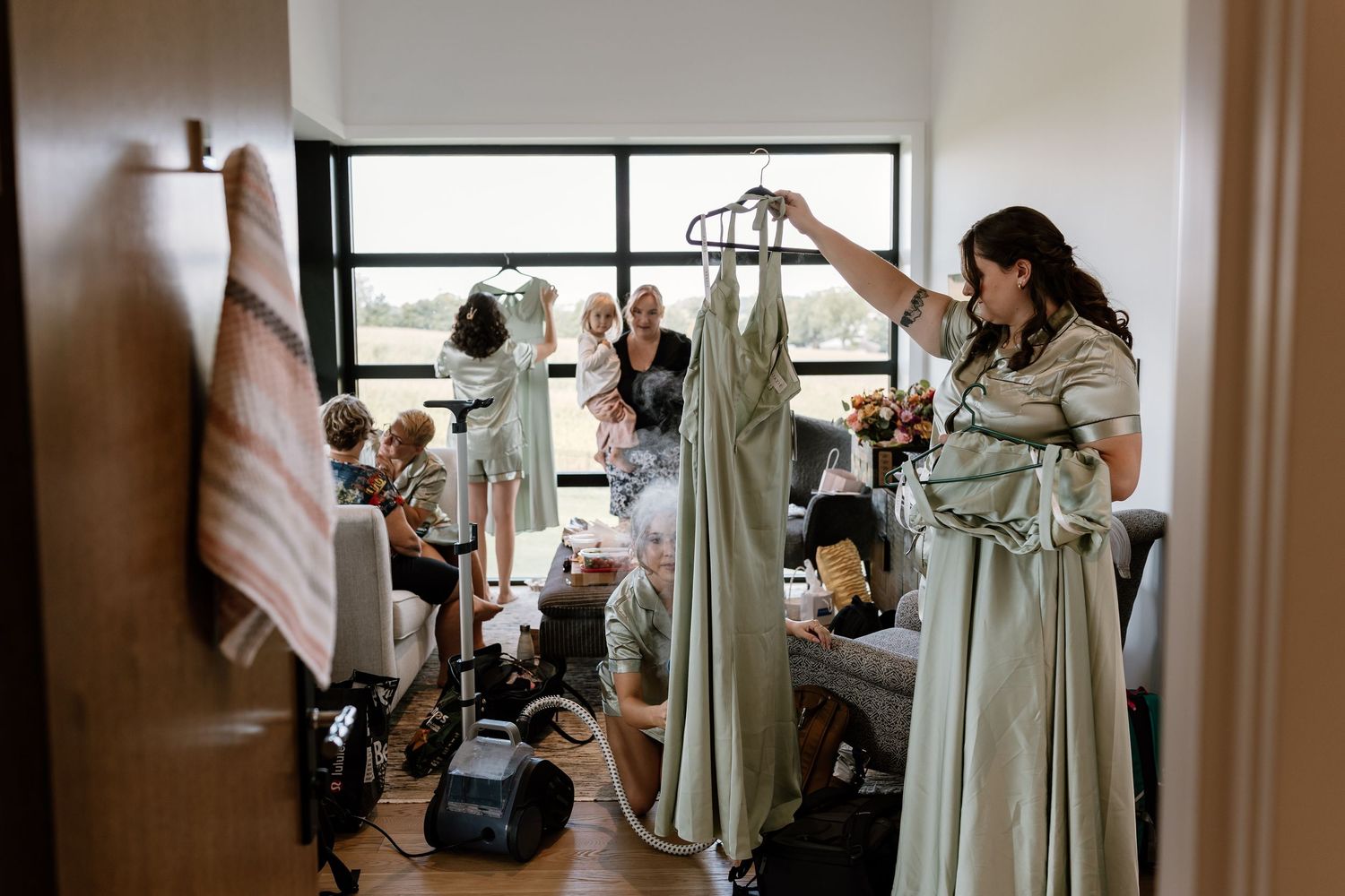 Bridesmaids getting ready in a room with sage green dresses hanging from a clothing rack.