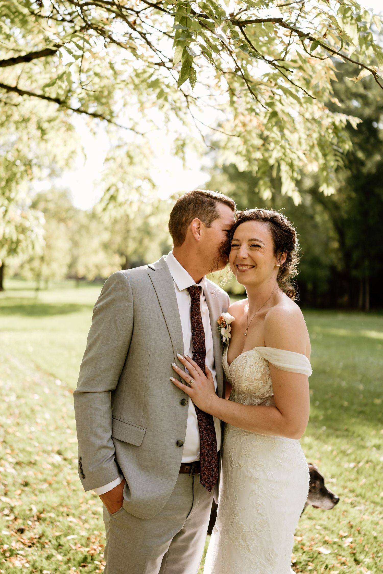 A joyful wedding moment captured under sunlit tree branches in a park setting.