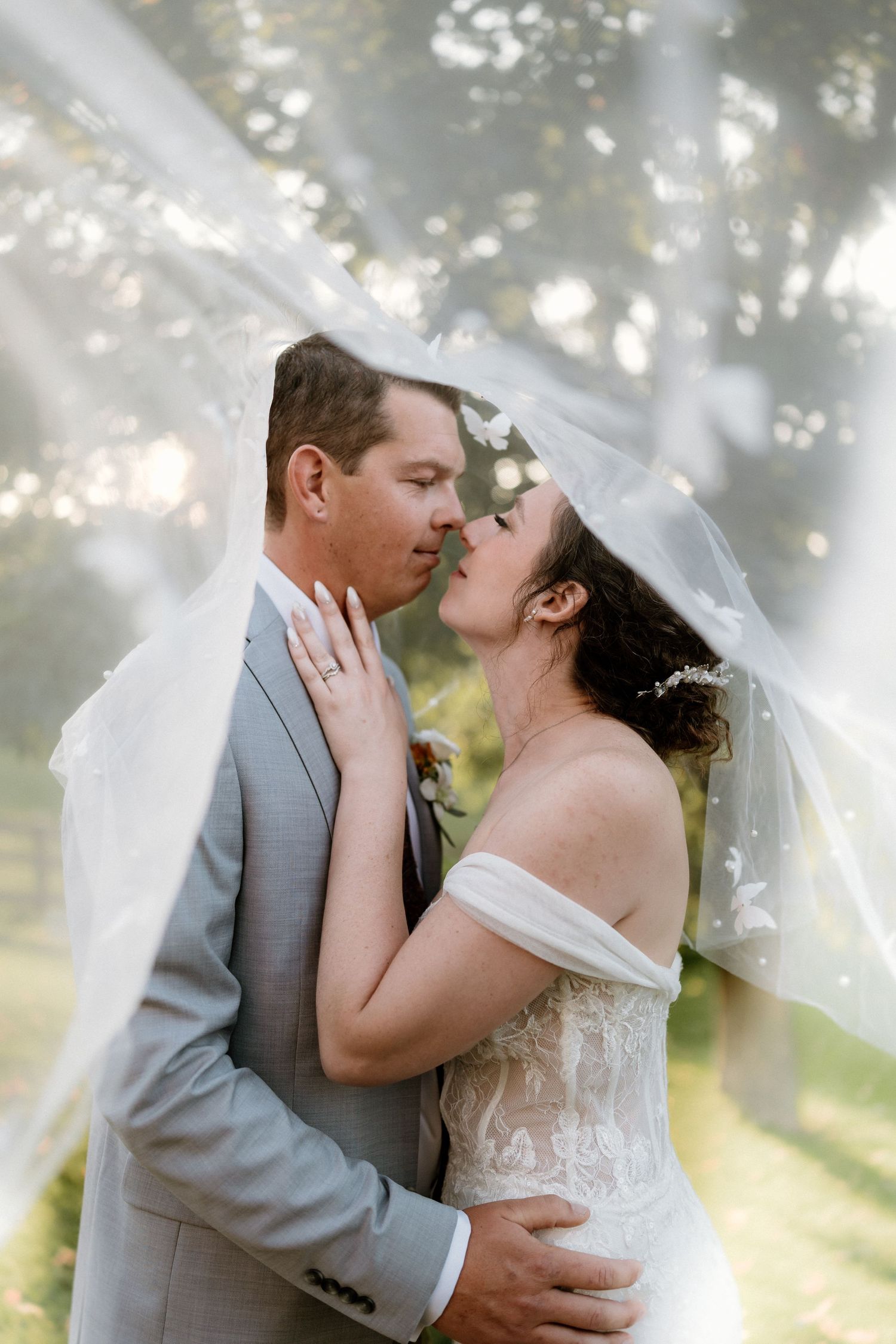 Bride and groom share an intimate kiss under flowing white veil in natural outdoor setting.