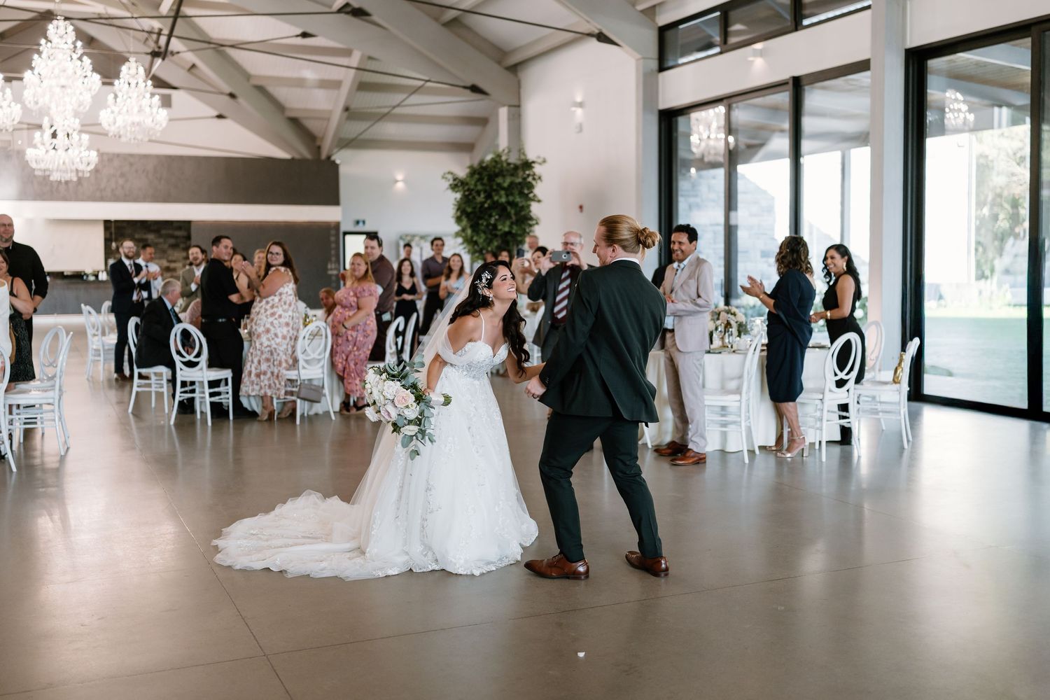 Bride and groom share first dance in elegant wedding venue with chandeliers and seated guests.
