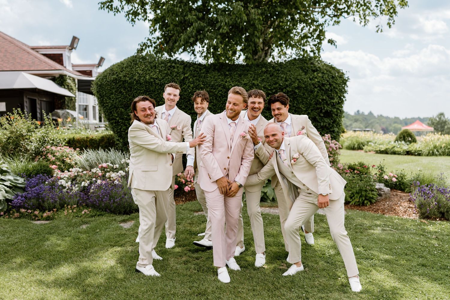 Groom in pink suit poses with groomsmen in tan suits on lush garden lawn at elegant outdoor wedding venue.