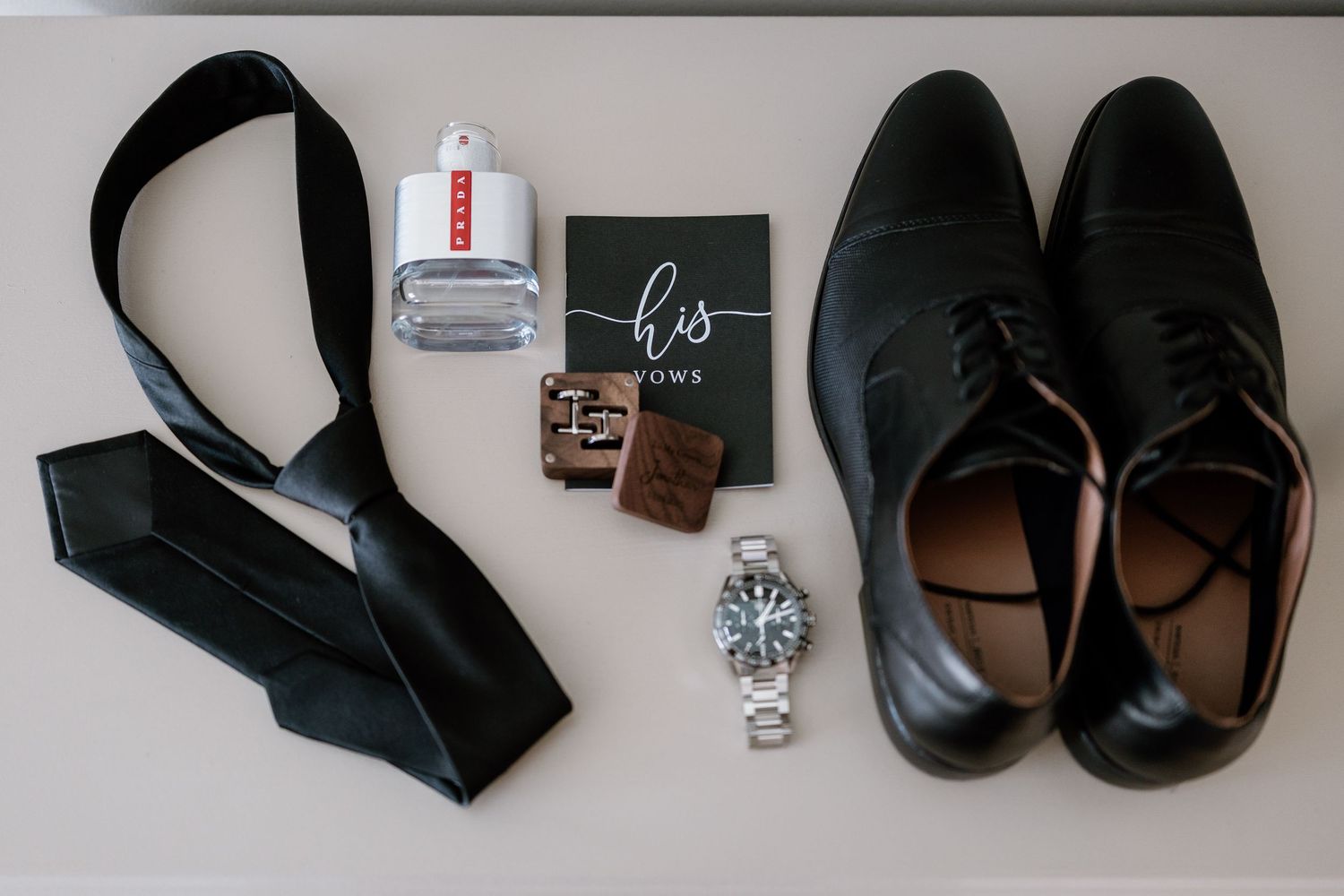 Wedding accessories including black dress shoes, tie, watch and rings laid flat on white surface.