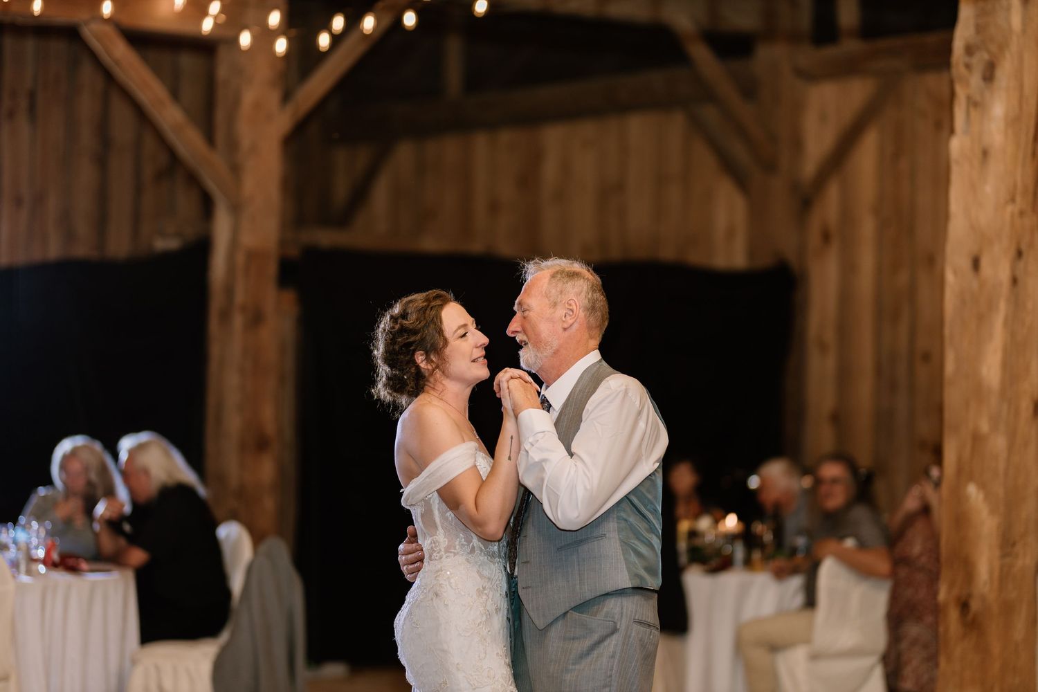Wedding reception dance in a rustic barn venue with wooden beams and string lights creating a warm atmosphere.