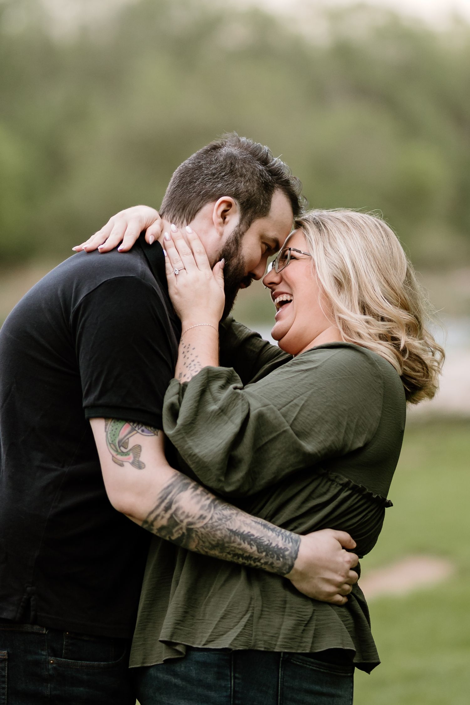 Couple embracing and laughing outdoors, man with tattoos in black shirt and woman in green top.