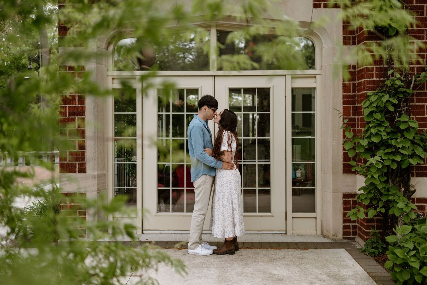 Couple embracing romantically in front of elegant French doors at a brick building surrounded by lush greenery.