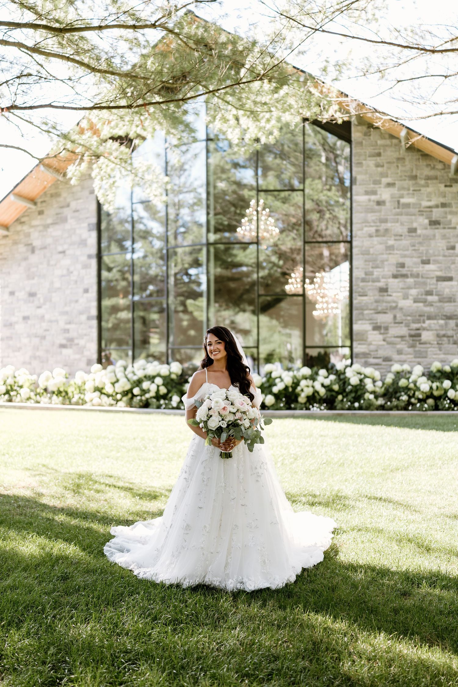 Bride in elegant off-shoulder ball gown holding bouquet stands on lawn before modern stone chapel.