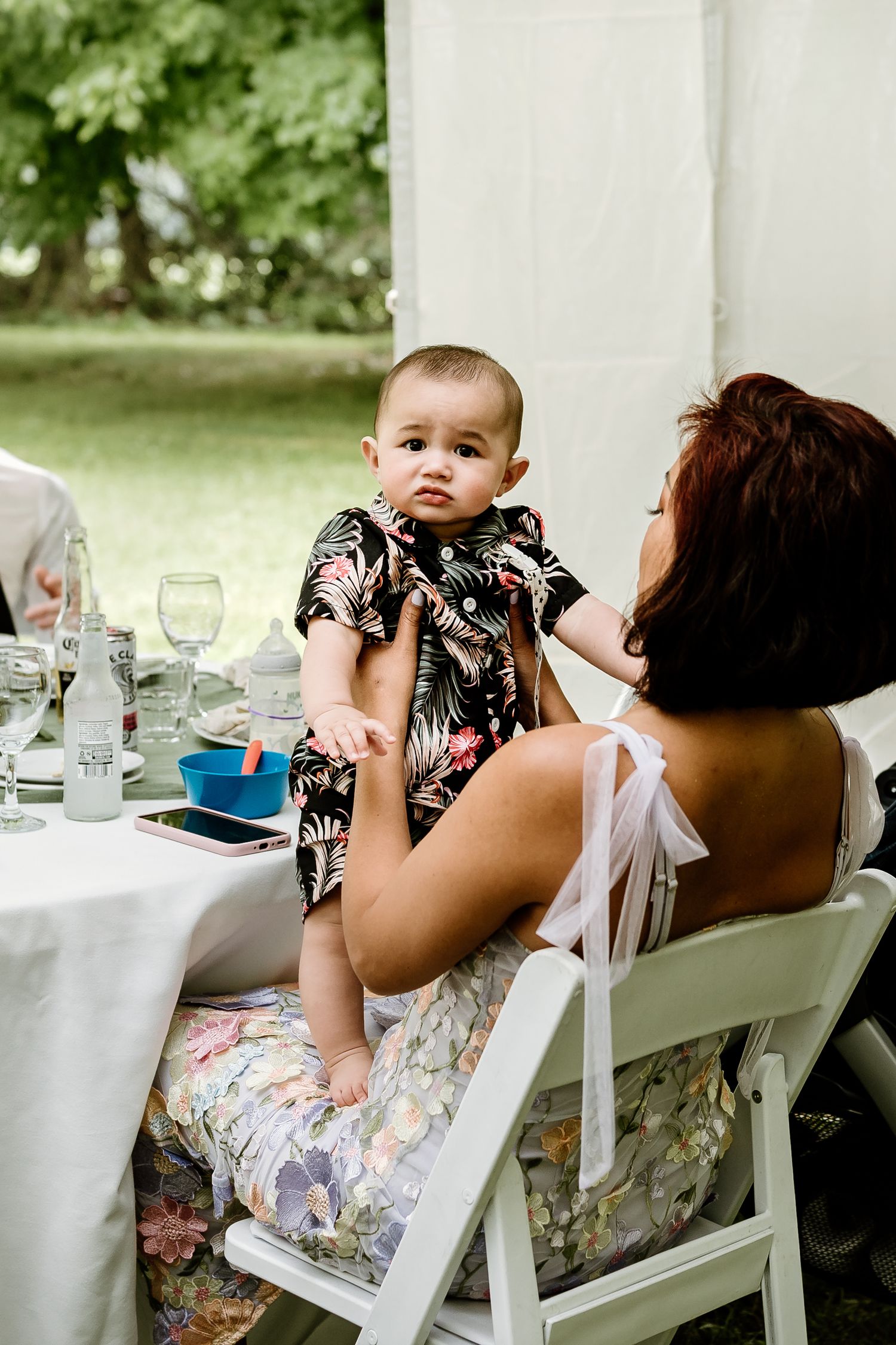 Guest holds baby wearing floral outfit at outdoor wedding reception.