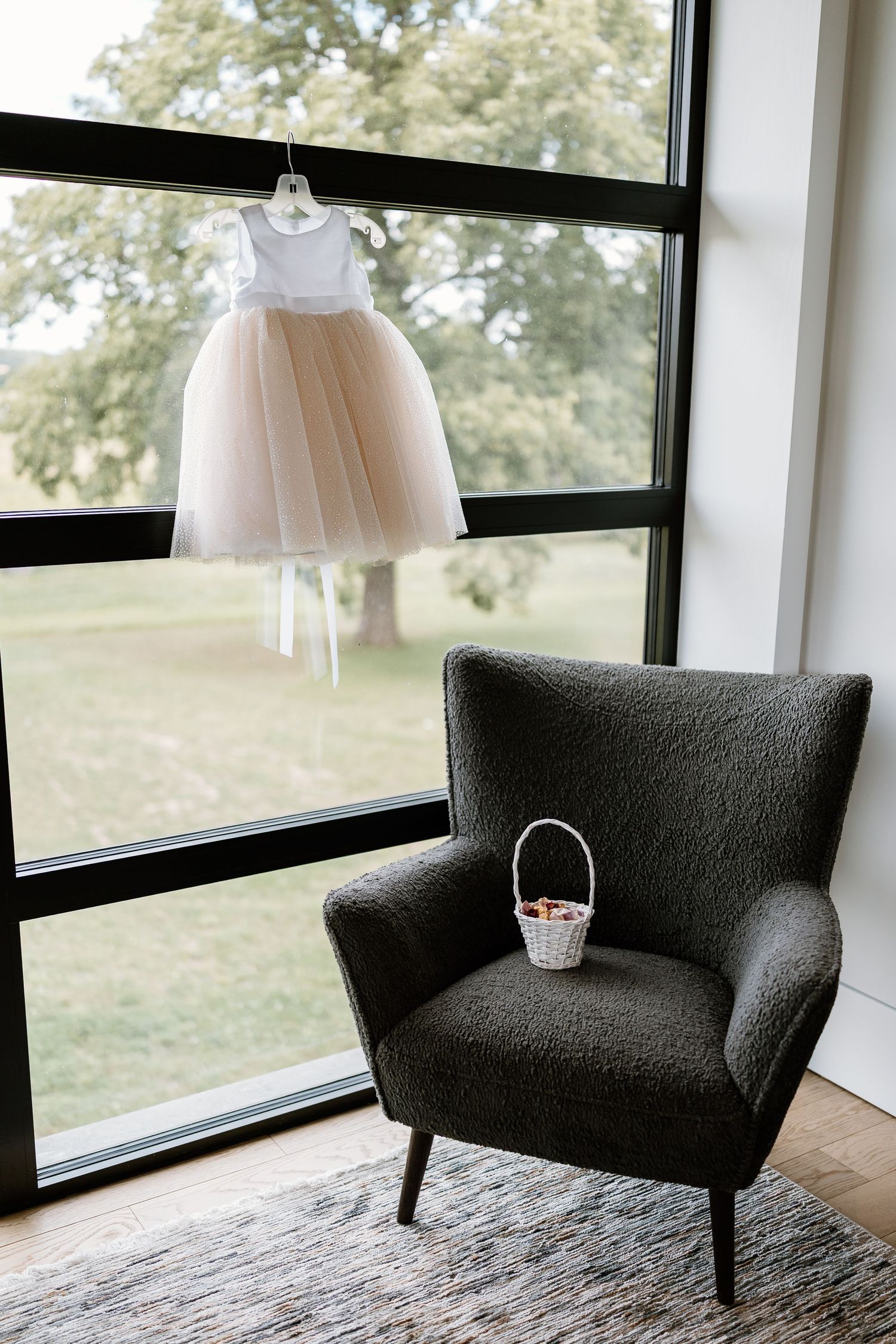 A delicate tulle flower girl dress hangs by a window next to a gray armchair with a small basket.