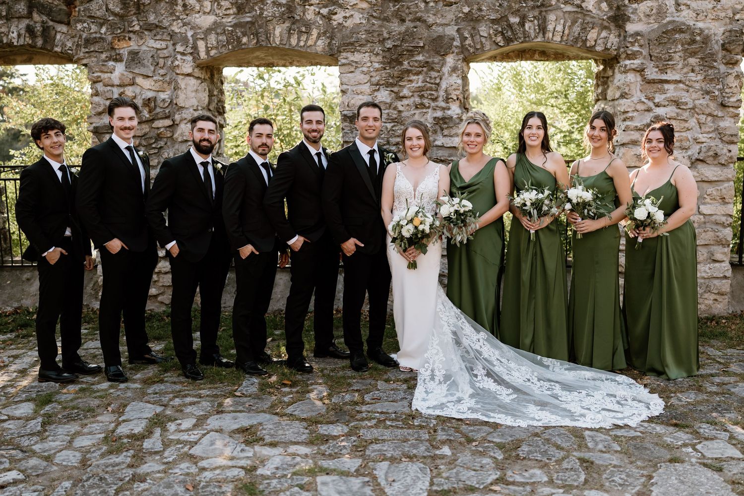 Wedding party poses against rustic stone wall with bridesmaids in olive green dresses.