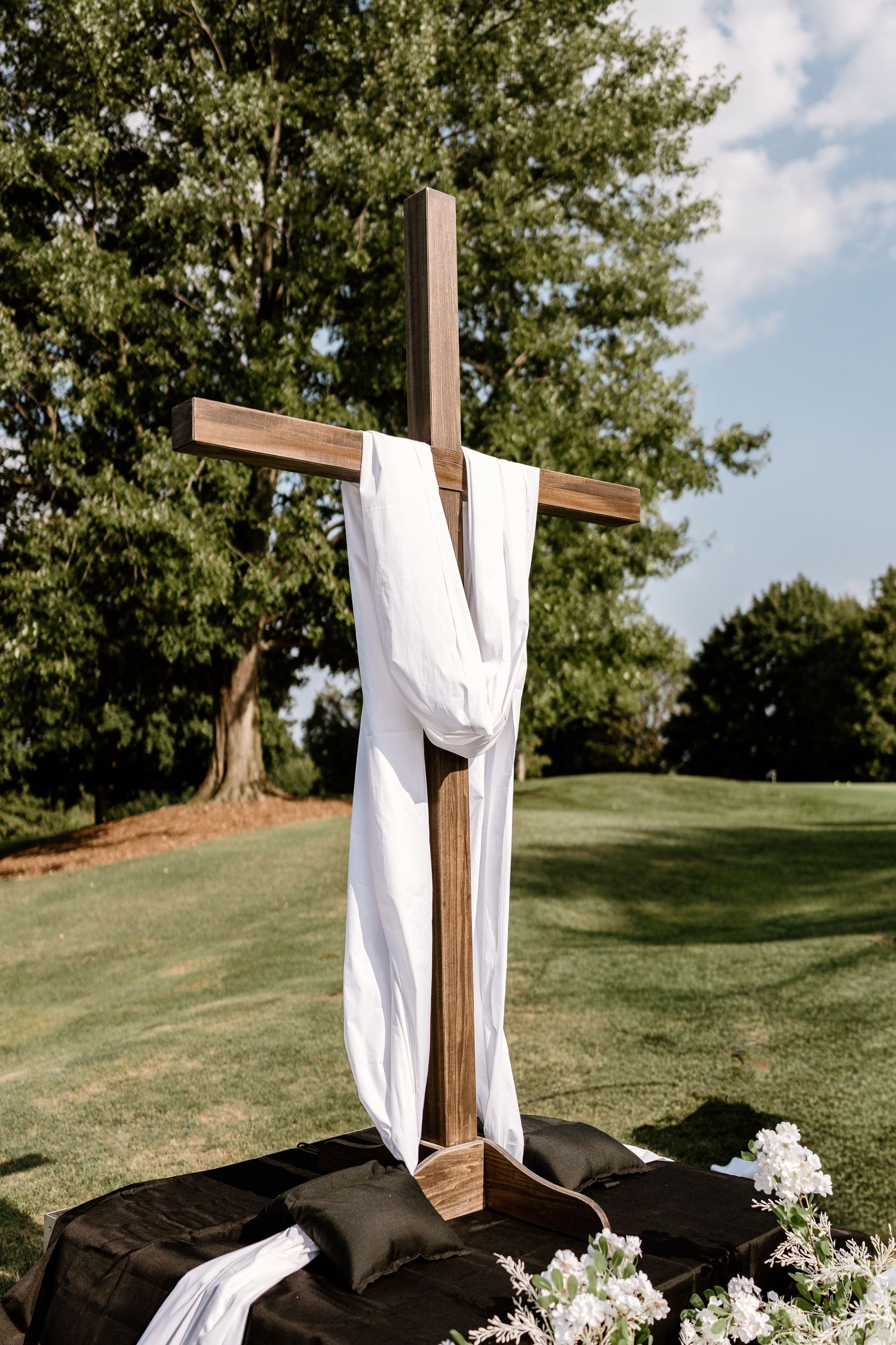 Rustic wooden cross draped with white fabric and floral arrangements for an outdoor wedding ceremony.