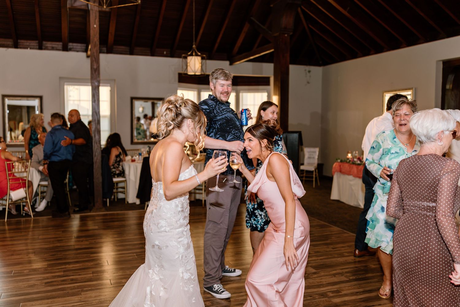 Bride in white gown dancing with guests in elegant rustic wedding reception venue with wood beam ceiling.