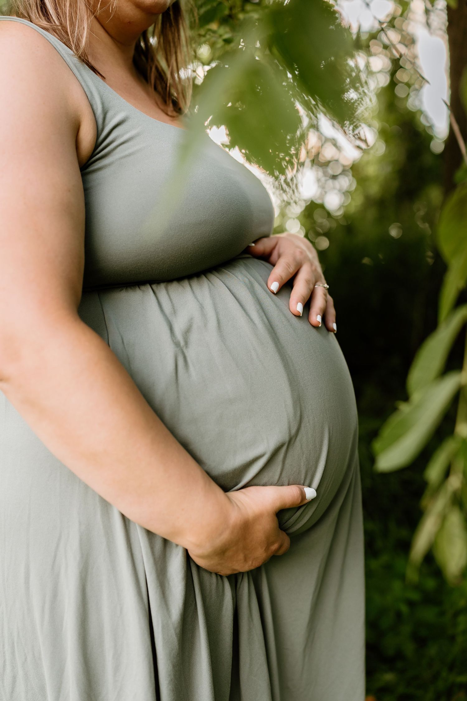 Pregnant woman in sage green maxi dress cradling baby bump outdoors with lush green foliage background.