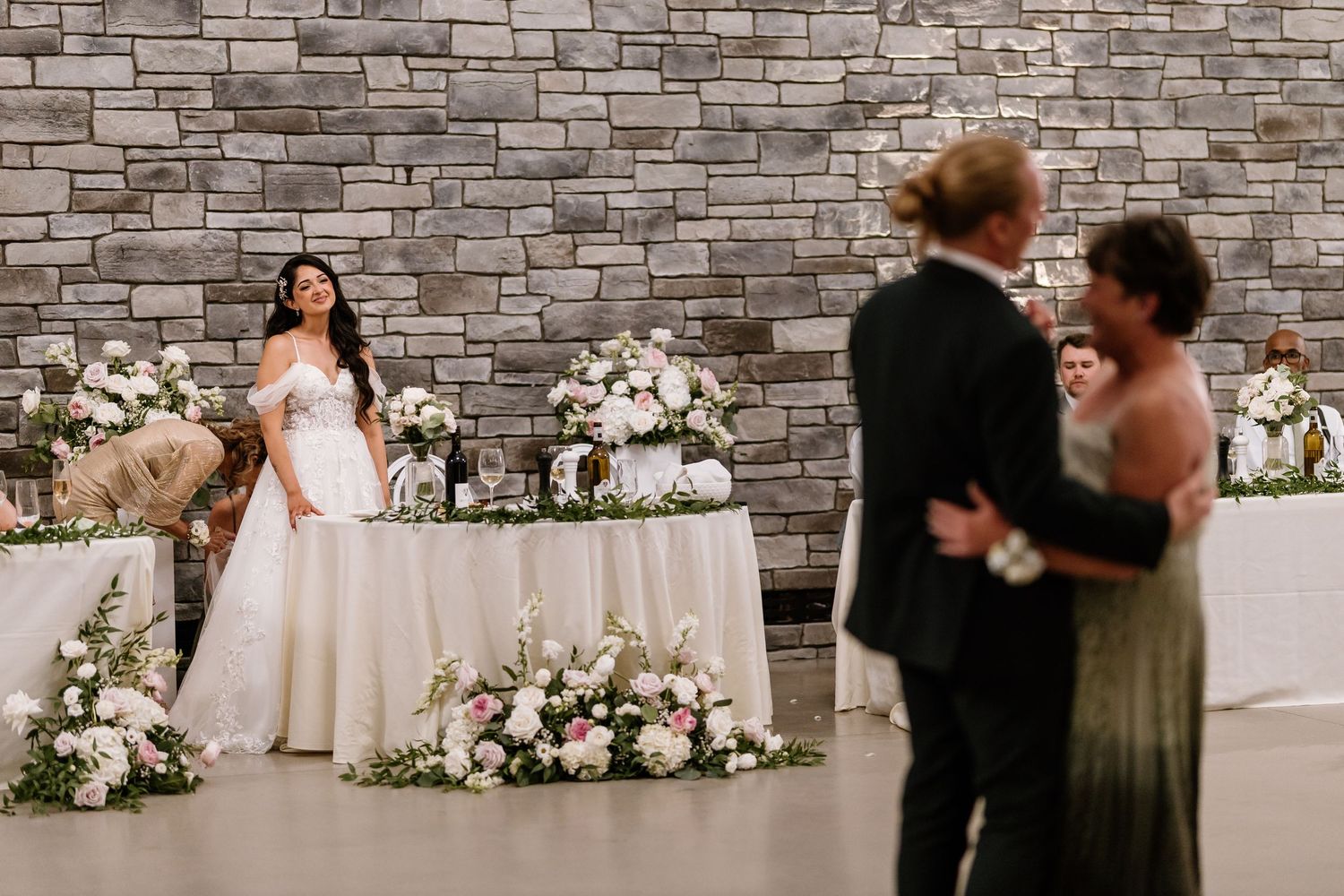 Bride stands by floral-decorated sweetheart table as couple dances in foreground at elegant stone-walled wedding reception.