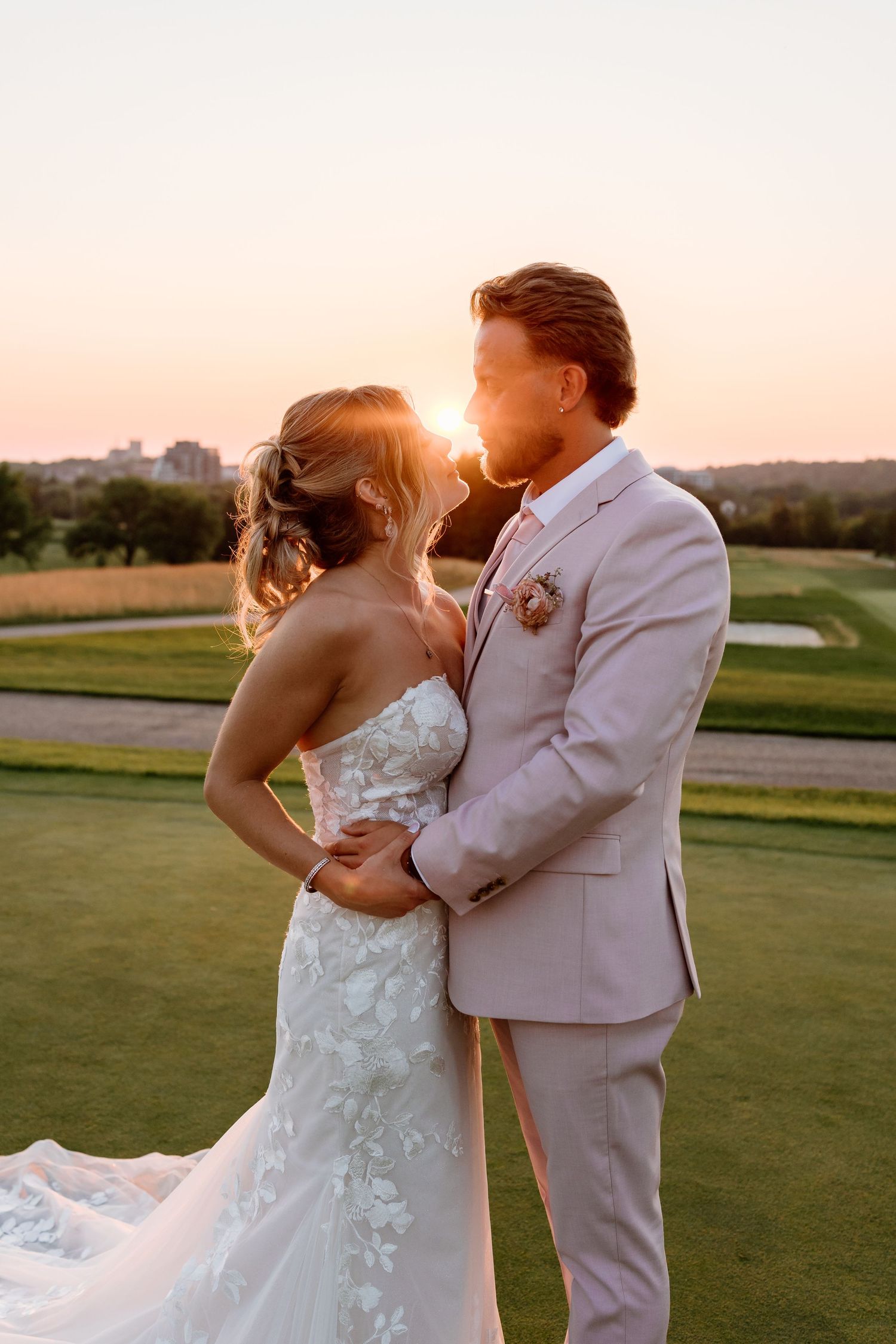 A bride and groom share a romantic moment at sunset on a golf course, the groom in a gray suit.